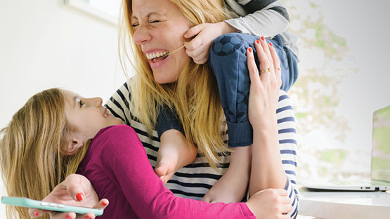 A mother laughing and playing with her two children while holding her phone