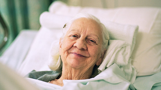 An elderly woman with white hair laying in a hospital bed and smiling.