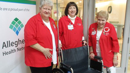 Three AHN volunteers wearing their red AHN volunteer coats standing around a wheelchair.