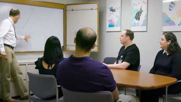 Four psychiatry residents sit around a conference table, listening to a lecture from faculty.