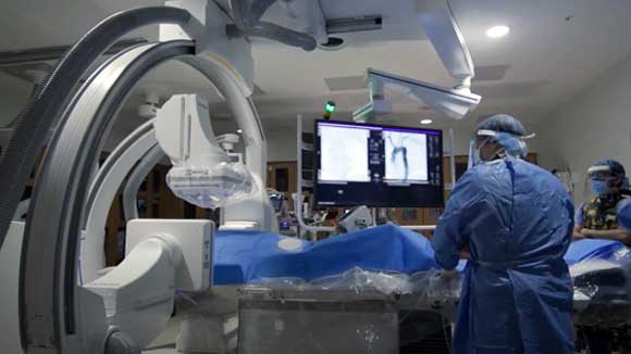 An interventional radiology resident performs a procedure in a surgical suite with large monitors.