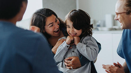 A happy child laughs while being held by their loving parent, with a medical professional smiling nearby.