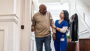 A nurse helping an older patient walk down a hallway while he holds on the wall for support