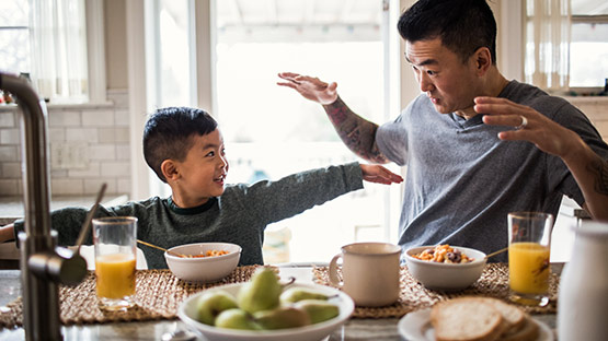 A father and son being playful as they eat breakfast, which consists of bowls of cereal and orange juice.