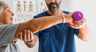 A medical professional in scrubs assists a senior woman with strength therapy exercises using light weights.