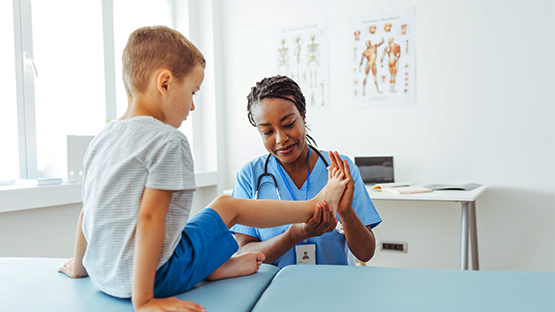 A medical professional in scrubs gently examines a young boy's foot during pediatric therapy.