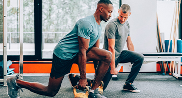 A medical professional guides a client through a sports therapy exercise using kettlebells in a gym.