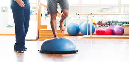 A male patient performs a balance exercise on a stability ball, guided by a medical professional in scrubs.